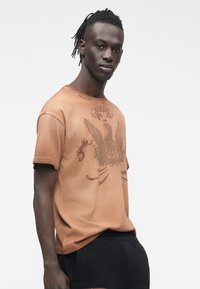 Hombre con cabello trenzado, lleva una camiseta marrón de Diesel con un gráfico de águila y pantalones negros, de pie frente a un fondo blanco liso.