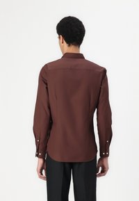 Cămașă lungă de culoare maro, cu mâneci lungi și guler, având o textură fină și mansete cu nasturi, purtată cu pantaloni negri, prezentată din spate.