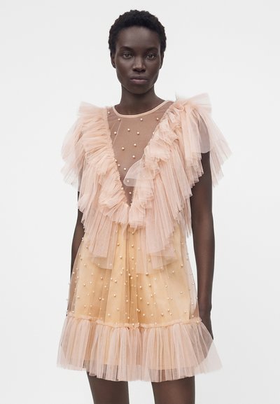 Γυναίκα με φόρεμα από ροδακινί τούλι με στρωματοποιημένες βολάν και μικρές πέρλες ως διακόσμηση, που στέκεται μπροστά σε απλό λευκό φόντο.