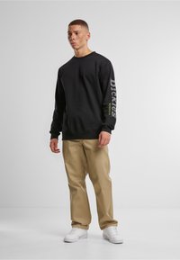 Fekete pulóver bordázott mandzsettákkal és szélekkel, a bal tenyéren "Dickies" felirattal fehér és zöld színben. Beig színű nadrág és fehér cipő egészíti ki a szettet.