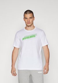 Valge lühikeste varrukatega T-särk, mille rinnale on trükitud roheline "HUMMEL" logo. Sile tekstuur, ümar kaelus, kokku sobitatud hallide trennisärkide pükstega.