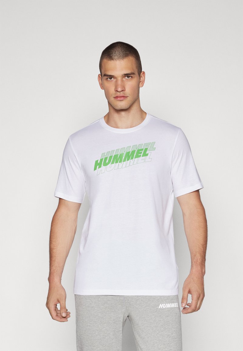 Valge lühikeste varrukatega T-särk, mille rinnale on trükitud roheline "HUMMEL" logo. Sile tekstuur, ümar kaelus, kokku sobitatud hallide trennisärkide pükstega.