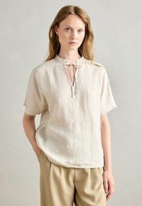 Beige top z lněné směsi s krátkým rukávem, stojáčkovým límcem a vázáním na výstřihu. Má jednoduchý, volný střih a jemné švy.