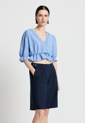 Donna in piedi con le mani nelle tasche di pantaloncini blu navy, indossa una blusa corta azzurro chiaro con maniche a sbuffo e scollo a V su sfondo semplice.