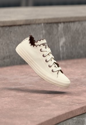 Zapatilla baja color blanco hueso con bordes en forma de ondas y ribete marrón flotando sobre escalones de piedra grises y rosas.