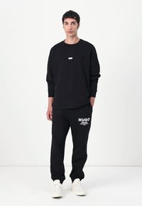 Black sweatshirt with a small logo; paired with black sweatpants featuring white "HUGO" text; model wearing white sneakers.