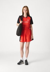 Červený športový dres s čiernymi rukávmi, elastické šortky a čierna taška. Na hrudi a na rukáve je logo. K tomu neformálna športová obuv.