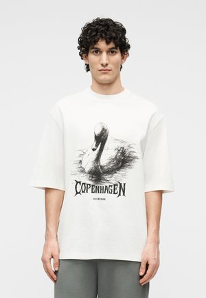 Νεαρός άνδρας με σγουρά μαλλιά που φοράει λευκό υπερμέγεθες T-shirt με μαύρο σκιασμένο κύκνο και τη λέξη "Κοπεγχάγη" σε στιλιζαρισμένη γραμματοσειρά.