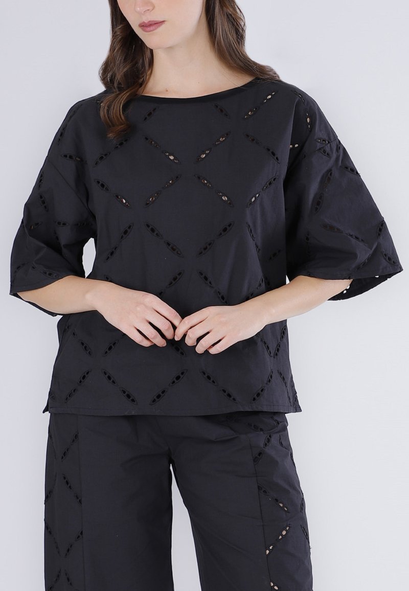 Femme portant un ensemble haut et pantalon noirs avec des motifs découpés en forme de losange, debout, les mains doucement jointes devant elle.