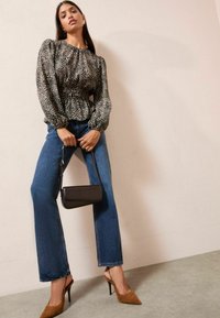 Blusa bianca e nera con pois e maniche a sbuffo, abbinata a jeans di denim blu a gambale svasato e décolleté a punta marroni. Tiene una piccola borsa nera.
