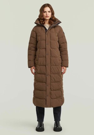 Frau trägt langen braunen gesteppte Wintermantel mit hohem Kragen, schwarze Hose und schwarze Stiefel, steht vor einem einfarbigen hellen Hintergrund.