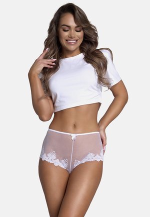 Lachende vrouw in een witte cropped t-shirt en doorzichtige hoge tailleonderbroek met kanten rand, poseert met haar hand bij haar gezicht.