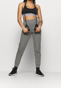 Sujetador deportivo negro con logo blanco, pantalones jogger grises ajustados, zapatillas negras y una cuerda para saltar en las manos, todo sobre un fondo liso.
