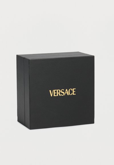 Μαύρο ορθογώνιο κουτί με χρυσό λογότυπο «VERSACE» στο κέντρο της μπροστινής πλευράς, τοποθετημένο σε απλό ανοιχτό γκρι φόντο.