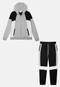 Grijze hoodie met rits, voorzien van zwarte en witte accenten, gecombineerd met zwarte en grijze broek met witte strepen en een trekkoord in de taille.