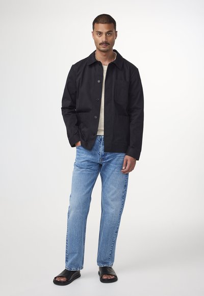 Zwarte katoenen overshirt met een puntige kraag, knoopsluiting aan de voorkant en borstzak, gecombineerd met lichtblauwe rechte jeans en zwarte sandalen.