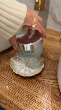 Lys blå krembeholder med sølvlokk holdt over en hvit blomsterformet tallerken med gullaksenter, plassert på en tremasse.