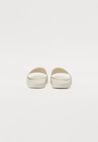 Sandale albe, ușoare, cu un design deschis, talpă plată și textură netedă. Au o formă minimalistă și o curea lată în partea de sus.