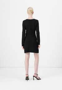 Black long-sleeve dress, fitted design, smooth texture, knee-length. Worn with strappy black heels featuring studded accents.