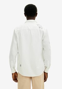 Camisa blanca de manga larga con puños abotonados, que presenta diseños impresos en negro en la parte posterior y en las mangas, confeccionada con una tela ligera.