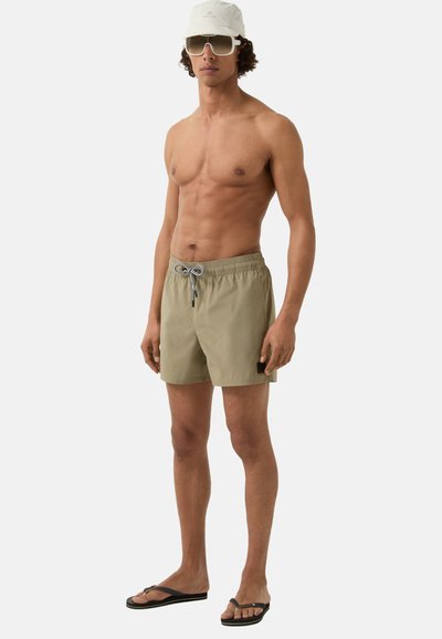 Khaki plavky s šňůrkou v pase, malou nášivkou, nošené s černými žabkami, bílou čepicí a oversized slunečními brýlemi.