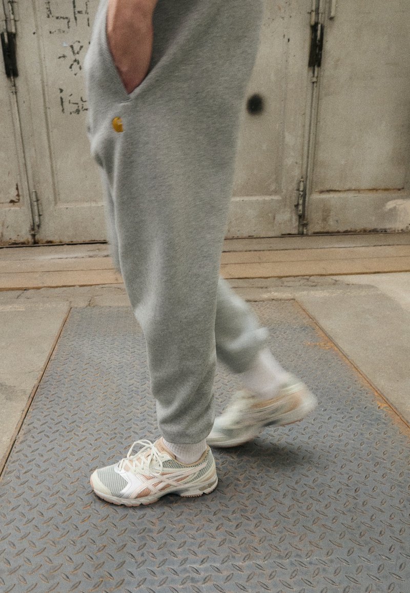 Person trägt graue Jogginghose und weiße Turnschuhe und geht auf einem strukturierten Metallboden in einer industriellen Innenumgebung.