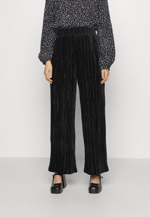 Kobieta ubrana w czarne plisowane szerokie spodnie z aksamitu, czarno-białą bluzkę w groszki oraz czarne buty Mary Jane na obcasie.