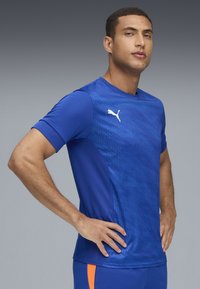 Blå sporttröja med en texturerad design och vertikala ränder, korta ärmar och en vit logotyp på bröstet. Andningsbart material.