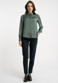 Zielona satynowa bluzka z kołnierzykiem i zapięciem na guziki, zestawiona z ciemnymi spodnimi w kratę. Materiał wydaje się gładki; krój dopasowany.
