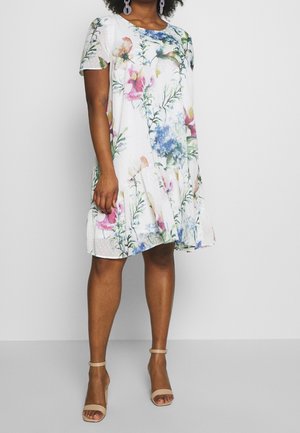 Frau trägt ein weißes knielanges Kleid mit bunten Blumenmustern und beigen Knöchelriemen-Pumps, steht vor einem schlichten hellen Hintergrund.