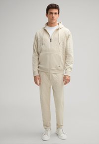 Felpa beige con cappuccio e pantaloni abbinati realizzati in tessuto testurizzato, caratterizzati da una zip frontale, cappuccio e maniche lunghe con vestibilità morbida. Sneakers bianche.
