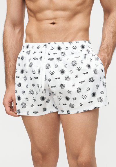 Hvite badeshorts med svarte sommerinspirerte trykk, inkludert soler, solbriller, fredstegn og «Moschino Swim»-logoer, brukt av en mannlig modell.
