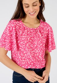 Blusa floreale rosa a maniche corte con scollo arricciato. Fiori bianchi stampati su tutto il tessuto, texture leggera e leggermente trasparente.