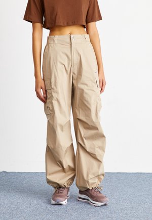 Person iført beige cargobukser med snøre i manchetterne, brun kort skjorte og mauve sneakers, stående på en grå tæppebelagt gulv.