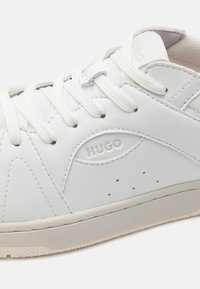 Baskets en cuir blanc avec un design à lacets, présentant un logo sur le côté, des perforations et une semelle en caoutchouc texturée.