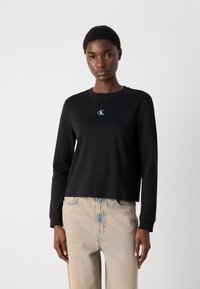 Schwarzes Sweatshirt mit langen Ärmeln und rundem Halsausschnitt, verziert mit einem kleinen tealfarbenen gestickten Logo auf der Brust, kombiniert mit hellblauen Jeans.