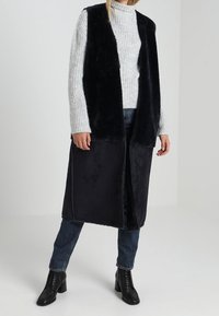 Černá vesta z umělé kožešiny s otevřeným předním dílem, nošená přes šedý žebrovaný rolák. Kombinováno s modrými džínami a černými šněrovacími botami. Hladká textura.