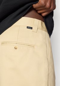 Krēmveida kokvilnas bikses ar klasisku piegriezumu, kas aprīkotas ar aizmugures kabatu ar pogu un mazu zīmola etiķeti tuvu jostai.