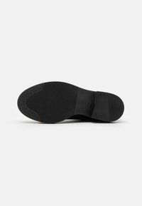 Semelle de chaussure noire avec surfaces antidérapantes texturées au niveau du talon, de la voûte plantaire et des orteils, présentée sur un fond blanc uni.