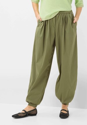 Person trägt lockere olivgrüne Hose mit elastischen Bündchen, hellgrünes Oberteil und schwarze Netz-Ballerinas, steht mit Händen in den Taschen.