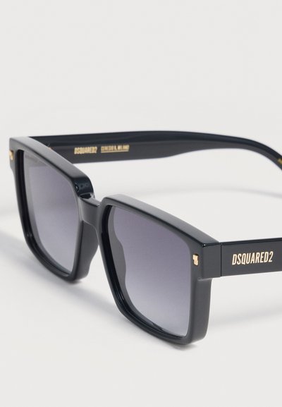Γυαλιά ηλίου με μαύρο σκελετό σε σχήμα τετράγωνο, φακούς με σκούρο βαθμιδωτό χρώμα, χρυσά στοιχεία και την επιγραφή "DSQUARED2" στα πλαινά μέρη.