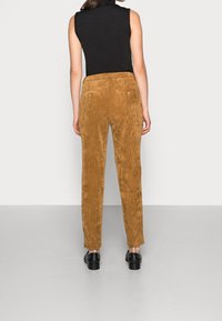 Femme portant un pantalon en velours côtelé couleur camel, un débardeur noir et des chaussures noires, debout face à un mur gris clair.