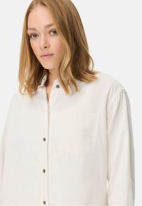Witte corduroy overhemd met een button-down kraag, voorzien van een enkele borstzak en bruine knopen langs de voorflap. Zachte textuur.