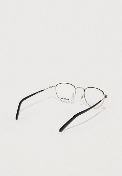 Ringikujulised metallist prillid õhukeste hõbedaste raamidega, mustad atsetaadist sangad ja läbipaistvad läätsed. Ühel läätsel on tuntud kaubamärgi logo.