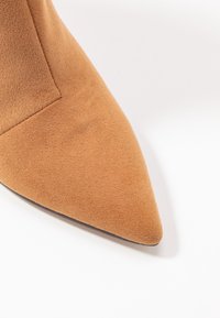 Pointe d'une chaussure en daim beige sur fond blanc, montrant les détails de la texture et des coutures.