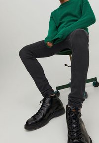 Suéter de punto verde con puños acanalados, jeans ajustados negros y botas negras con cordones, sobre una silla verde con ruedas contra un fondo claro.