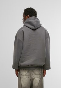 Szary, oversized hoodie z kapturem na sznurek, o miękkiej teksturze, szerokimi rękawami i ściągaczem u dołu, w połączeniu z jasnoszarymi spodniami z wyraźnymi szwami.