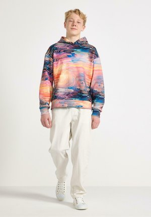 Teenagedreng iført hættetrøje med flerfarvet abstrakt mønster, cremefarvede bukser og hvide sneakers, gående mod en ensfarvet lys baggrund.