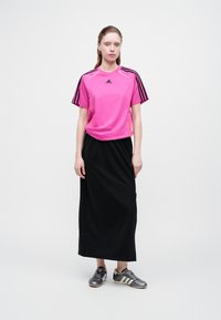 Žena stojí oblečená v ružovom športovom tričku Adidas, čiernej dlhé sukni a šedých teniskách Adidas na čistom bielom pozadí.