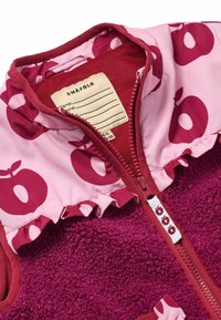 Fleecejacka med en djup rosa kropp och ljusrosa krage som har äppelmönster. Texturerad utsida och volang nertill med en dragkedja fram.